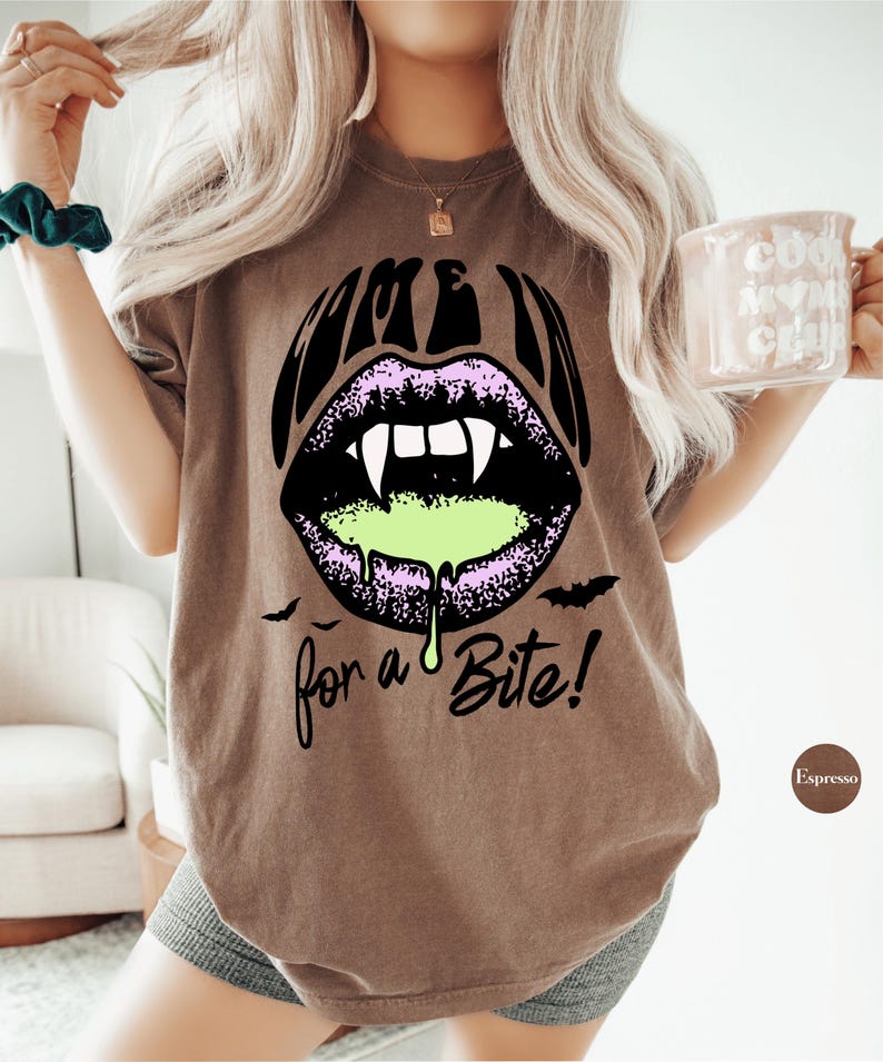 May include: A brown short-sleeve t-shirt featuring a graphic of purple lips with white fangs and green slime dripping from the mouth. The text "COME IN" is printed above the lips and "for a Bite!" is printed below, accompanied by two small black bats.