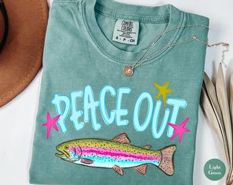 May include: A light green short-sleeve t-shirt with a crew neckline. The shirt features the text "PEACE OUT" in light blue letters, accented by two pink stars and one yellow star. Below the text is a colorful illustration of a rainbow trout fish.