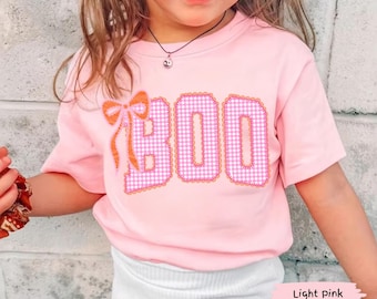 Puede incluir: Una camiseta de manga corta de color rosa claro con la palabra BOO en un patrón de cuadros vichy rosa y blanco con un borde festoneado naranja. Un lazo de cuadros vichy naranja está unido al lado izquierdo de la letra B.