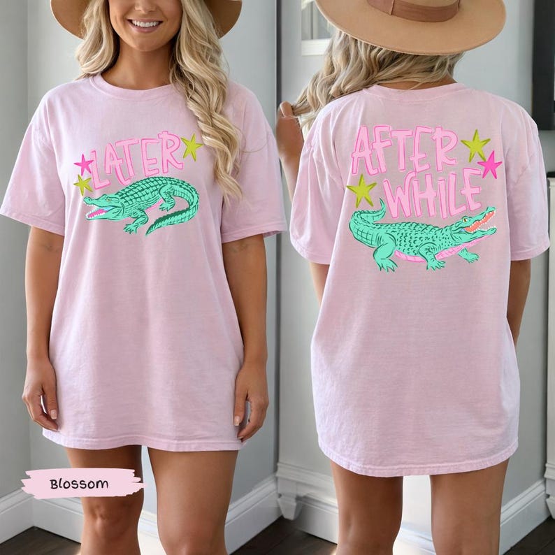 May include: A light pink t-shirt with a green alligator graphic and the word "LATER" on the front. The back of the shirt features the words "AFTER WHILE" and a matching alligator design. The shirt has short sleeves.