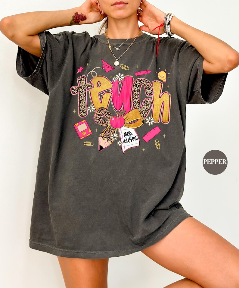 May include: Dark gray t-shirt with a colorful "Teach" graphic. The design includes a leopard print cross, apple, pencil, and school supplies. The shirt has a relaxed fit and is suitable for teachers.