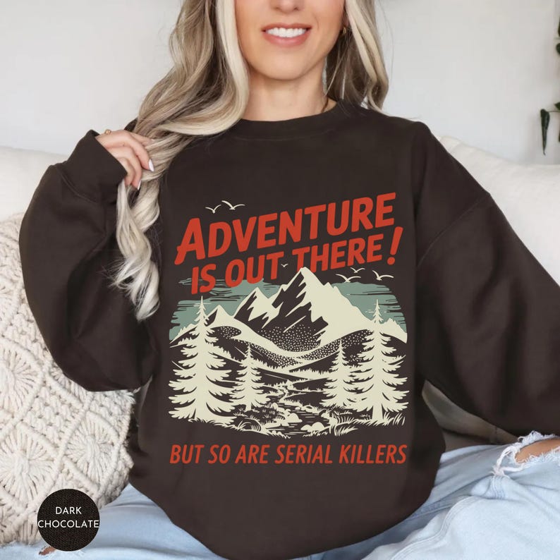 K&ouml;nnte beinhalten: Dunkelbraunes Sweatshirt mit Berg- und Waldgrafik. Der Text "ADVENTURE IS OUT THERE! BUT SO ARE SERIAL KILLERS" ist in Rot und Wei&szlig; gedruckt. Das Sweatshirt hat einen Rundhalsausschnitt und lange &Auml;rmel.
