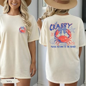 Puede incluir: Camiseta de color marfil con un dise&ntilde;o de cangrejo en la parte delantera y trasera. La parte delantera presenta un peque&ntilde;o dise&ntilde;o de cangrejo, mientras que la parte trasera muestra un cangrejo m&aacute;s grande con el texto "If Crabby, Please Return to the Beach".
