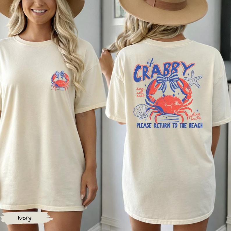Puede incluir: Camiseta de color marfil con un dise&ntilde;o de cangrejo en la parte delantera y trasera. La parte delantera presenta un peque&ntilde;o dise&ntilde;o de cangrejo, mientras que la parte trasera muestra un cangrejo m&aacute;s grande con el texto "If Crabby, Please Return to the Beach".