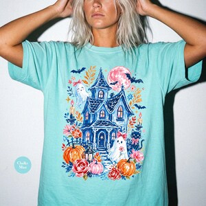 K&ouml;nnte beinhalten: Ein mintgr&uuml;nes T-Shirt mit einem farbenfrohen Grafikdruck eines dunkelblauen Spukhauses, zwei wei&szlig;en Geistern mit rosa Schleifen, orangefarbenen K&uuml;rbissen, roten und rosa Blumen, einer schwarzen Katze, Flederm&auml;usen und einem gro&szlig;en rosa Mond.