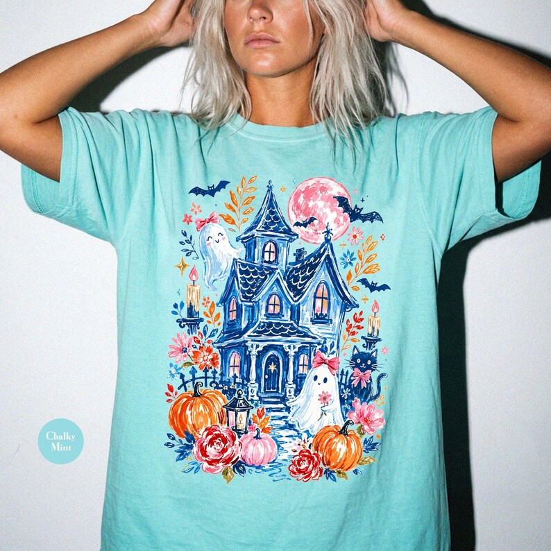 K&ouml;nnte beinhalten: Ein mintgr&uuml;nes T-Shirt mit einem farbenfrohen Grafikdruck eines dunkelblauen Spukhauses, zwei wei&szlig;en Geistern mit rosa Schleifen, orangefarbenen K&uuml;rbissen, roten und rosa Blumen, einer schwarzen Katze, Flederm&auml;usen und einem gro&szlig;en rosa Mond.