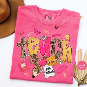 May include: A pink t-shirt with the word "TEACH" in gold and leopard print. The design includes an apple, pencil, notebook, and other school-themed graphics. The shirt has the text "Mrs. Wilson" on a notepad.