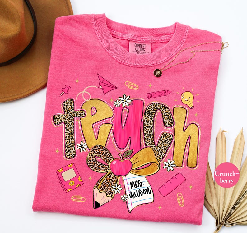 May include: A pink t-shirt with the word "TEACH" in gold and leopard print. The design includes an apple, pencil, notebook, and other school-themed graphics. The shirt has the text "Mrs. Wilson" on a notepad.