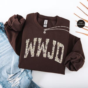 May include: A dark chocolate-colored sweatshirt with the letters "WWJD" in a camouflage pattern. A silver necklace with a pendant is on the sweatshirt. The sweatshirt is paired with blue jeans.
