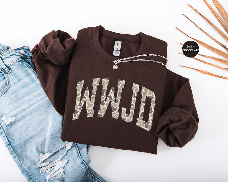 May include: A dark chocolate-colored sweatshirt with the letters "WWJD" in a camouflage pattern. A silver necklace with a pendant is on the sweatshirt. The sweatshirt is paired with blue jeans.