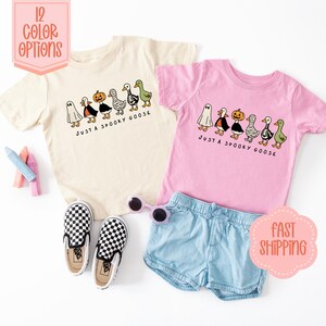 May include: Two children's t-shirts, one cream and one pink, with a Halloween goose design. The shirts feature cartoon geese dressed as a ghost, a witch, a pumpkin, and a mummy. Text reads "JUST A SPOOKY GOOSE".