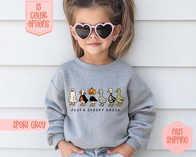 May include: A grey sweatshirt with a Halloween-themed design. It features cartoon geese in costumes and the phrase "JUST A SPOOKY GOOSE". The image also includes text saying "13 COLOR OPTIONS" and "SPORT GREY".