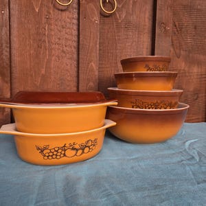 May include: A set of amber and brown glass kitchenware including two lidded casserole dishes with handles and three nesting mixing bowls. Each piece features a black line art illustration of grapes and apples around the exterior.