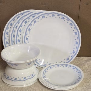 Può includere: Un servizio di piatti bianco con un motivo floreale blu. Il set include diversi piatti grandi, piatti piccoli e ciotole. I piatti e le ciotole hanno un bordo floreale blu. La tavola è apparecchiata su una tovaglia di pizzo bianco.