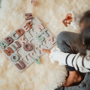 以下が含まれることがあります： 子供が木製のアルファベットパズルで遊んでいます。パズルは薄い灰色のベースに、各文字の個別のスロットがあります。文字はピンク、茶色、緑の様々な色合いです。子供は「R」の文字を手に持っています。
