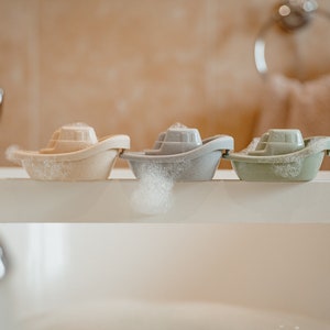 Puede incluir: Tres juguetes de baño con forma de barcos en una bañera. Los barcos son beige, gris y verde. Los barcos flotan en el agua y hay burbujas alrededor de ellos.