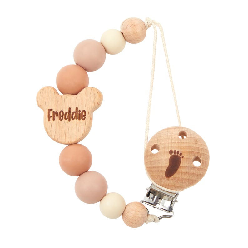 May include: A wooden pacifier clip with the name "Freddie" engraved on a bear-shaped charm. The clip features pastel-coloured beads and a round wooden piece with a footprint design. A metal clip is attached for securing.