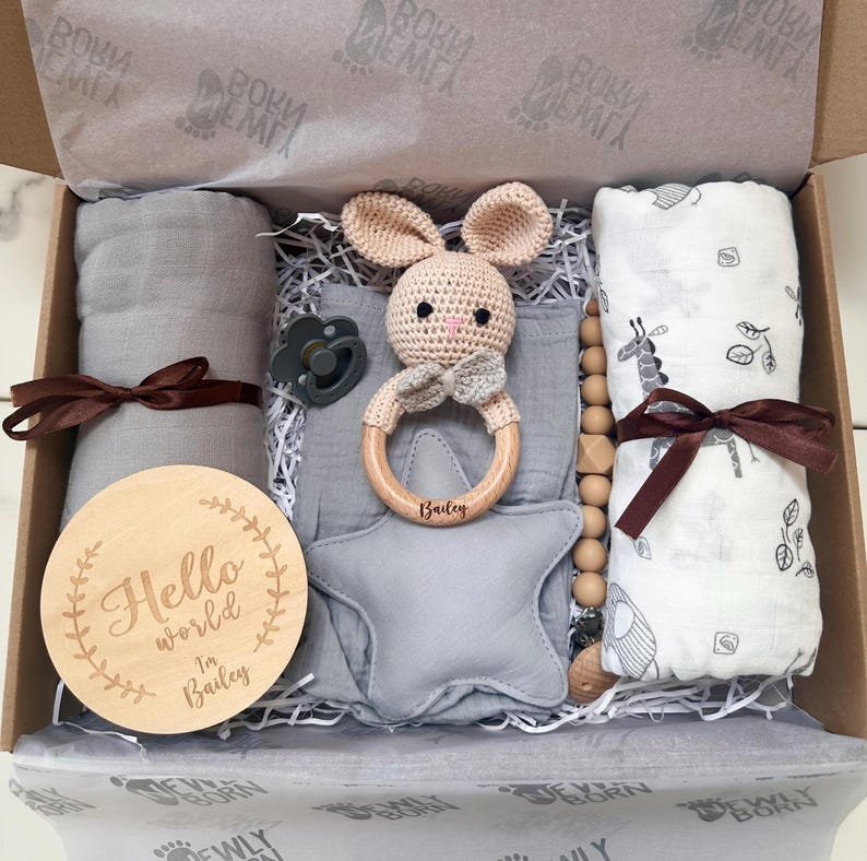 May include: A baby gift box filled with a gray swaddle blanket, a wooden teether, a pacifier, a star-shaped pillow, and a bunny-shaped rattle. The wooden disc reads "Hello world I'm Bailey". The box is lined with tissue paper.