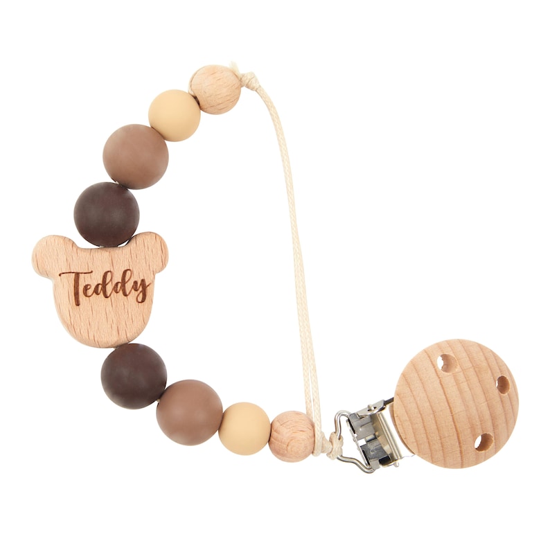 May include: A baby's dummy clip featuring a wooden teddy bear charm engraved with the name "Teddy". The clip has a series of brown, tan, and beige beads, and a wooden round dummy holder with three holes.