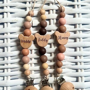 May include: Three wooden baby pacifier clips with neutral-toned beads. Each clip has a wooden bear-shaped charm with a name: "Freddie", "Teddy", and "Honey". The clips have a wooden base with a metal clasp.