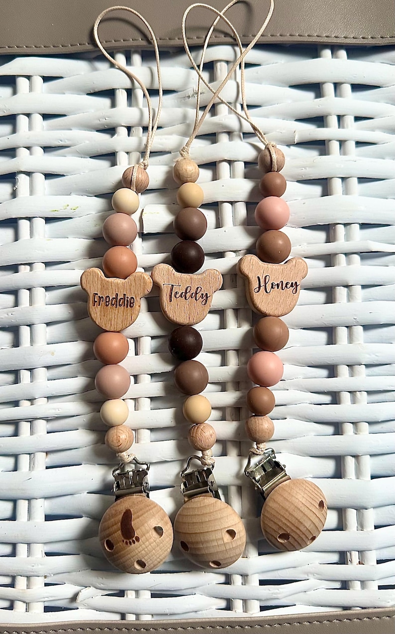 May include: Three wooden baby pacifier clips with neutral-toned beads. Each clip has a wooden bear-shaped charm with a name: "Freddie", "Teddy", and "Honey". The clips have a wooden base with a metal clasp.