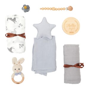 May include: A collection of baby items, including a pacifier, wooden bead clip, swaddling blankets, a star-shaped pillow, a wooden sign with text, and a crochet bunny rattle. The blankets are tied with brown ribbons.