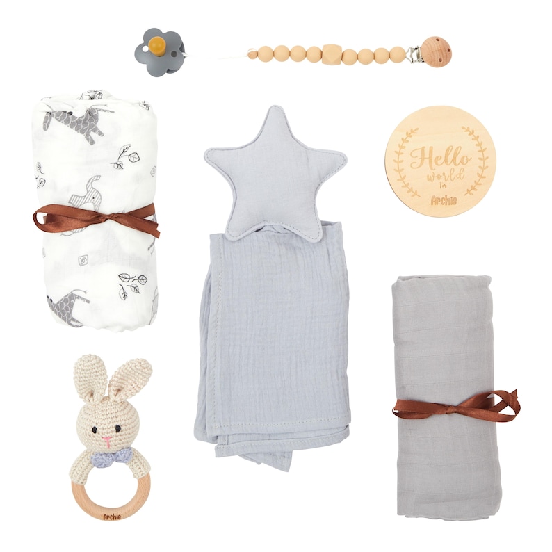 May include: A collection of baby items, including a pacifier, wooden bead clip, swaddling blankets, a star-shaped pillow, a wooden sign with text, and a crochet bunny rattle. The blankets are tied with brown ribbons.