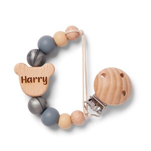 May include: A wooden baby pacifier clip with the name "Harry" engraved on a bear-shaped wooden piece. The clip features a series of grey, beige, and wooden beads. The pacifier clip is attached to a wooden round pacifier holder.