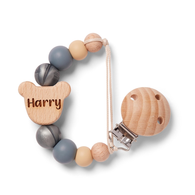 May include: A wooden baby pacifier clip with the name "Harry" engraved on a bear-shaped wooden piece. The clip features a series of grey, beige, and wooden beads. The pacifier clip is attached to a wooden round pacifier holder.