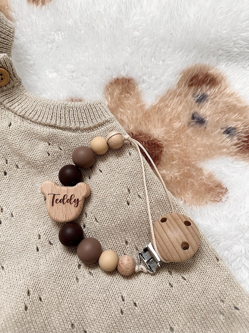 May include: A wooden baby pacifier clip with the name "Teddy" engraved on a bear-shaped charm. The clip features brown, beige, and wooden beads. The pacifier clip is attached to a wooden pacifier holder.