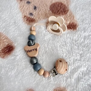 May include: A cream-coloured baby's dummy and wooden bead clip. The clip has the name "Harry" engraved on a wooden bear-shaped piece. The beads are in shades of grey, beige, and wood. The clip is silver.
