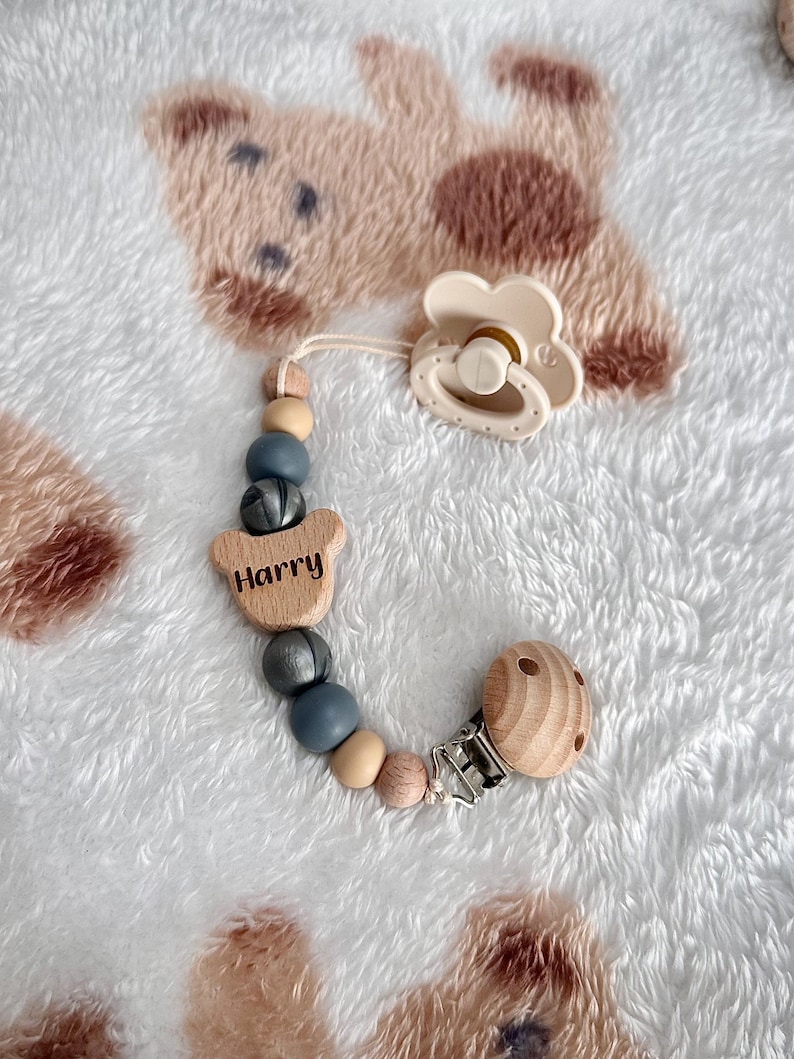 May include: A cream-coloured baby's dummy and wooden bead clip. The clip has the name "Harry" engraved on a wooden bear-shaped piece. The beads are in shades of grey, beige, and wood. The clip is silver.