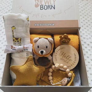 May include: A gift box for a newborn baby, filled with baby essentials. Includes a giraffe-printed blanket, a crochet lion rattle, a star-shaped plush, a pacifier, a wooden teether, and a wooden sign that says "Hello world".