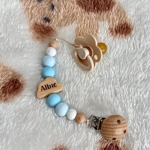 May include: A beige baby pacifier with a flower-shaped shield and a wooden bead clip. The pacifier is attached to a beaded chain with blue, white, and wooden beads. A wooden cloud charm with the name "Albie" is also included.