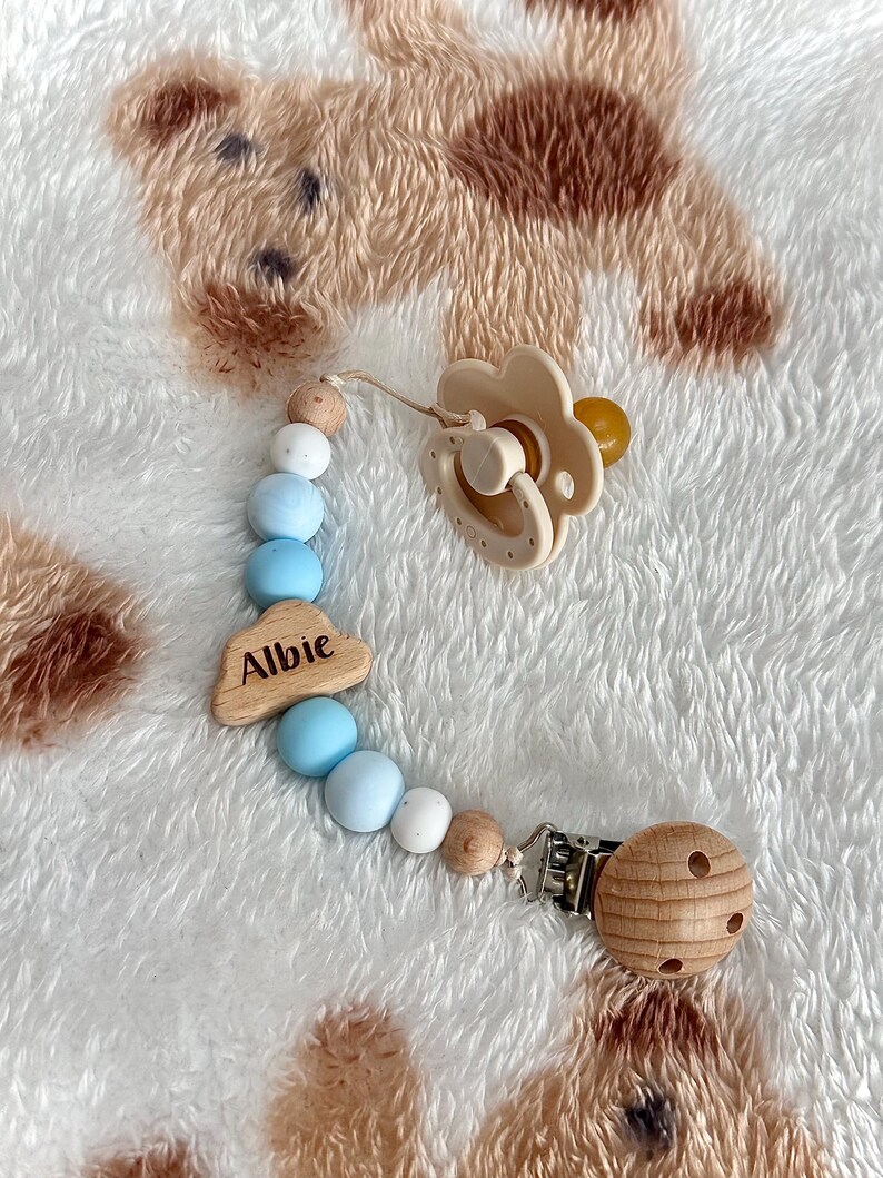 May include: A beige baby pacifier with a flower-shaped shield and a wooden bead clip. The pacifier is attached to a beaded chain with blue, white, and wooden beads. A wooden cloud charm with the name "Albie" is also included.
