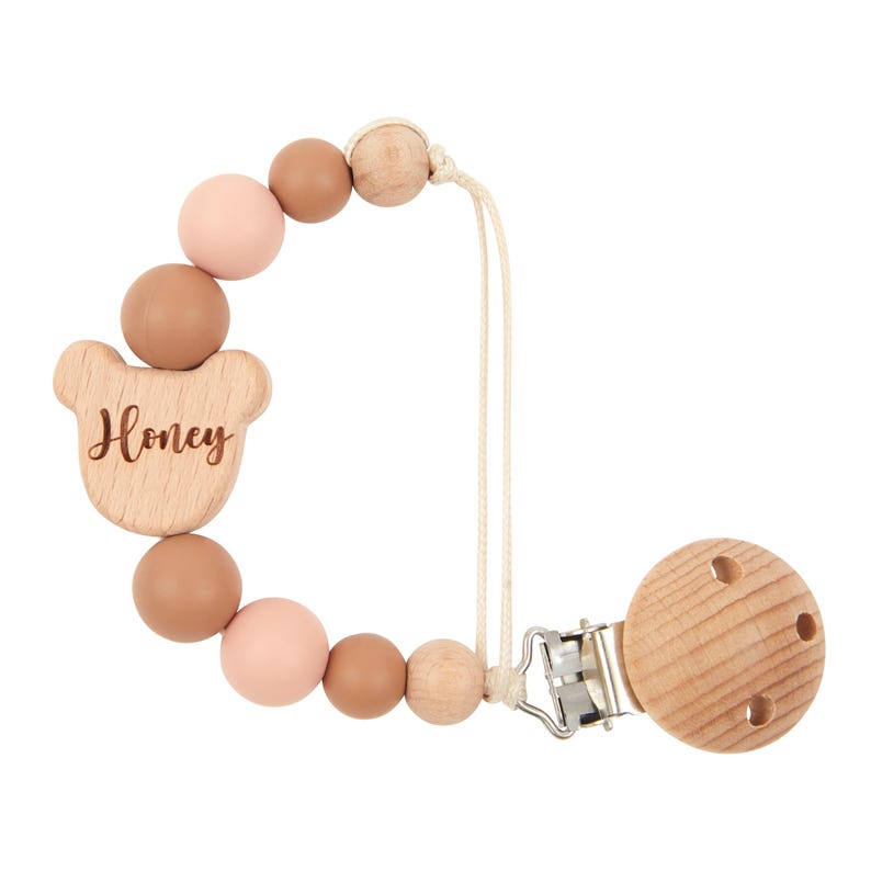 May include: A wooden pacifier clip with a bear-shaped charm that says "Honey". The clip features round beads in pink, brown, and natural wood. The clip has a wooden disc with three holes.