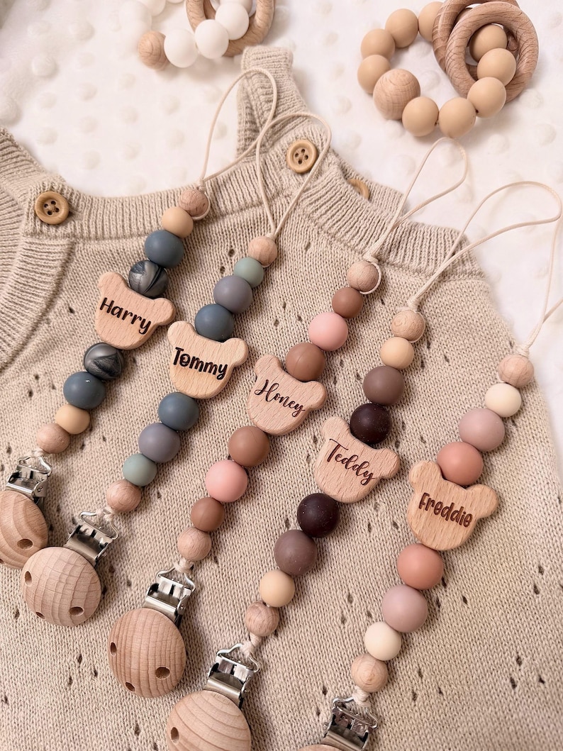 May include: Baby pacifier clips with names "Harry", "Tommy", "Honey", "Teddy", and "Freddie" engraved on bear-shaped wooden pieces. The clips feature a variety of coloured beads and wooden pacifier holders. A wooden teething ring and a beaded bracelet are also visible.