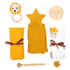 May include: A collection of baby items in shades of yellow and white. Includes a crochet lion rattle, a star-shaped pillow, a muslin blanket, a swaddling blanket with a fox print, a wooden pacifier clip, and a wooden disc with the text "Hello world in Alex."
