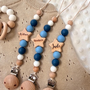 Puede incluir: Pinzas para chupete de bebé de madera con etiquetas con nombres en forma de estrella. Las pinzas presentan una mezcla de cuentas azules, blancas y de madera. Cada estrella está grabada con un nombre: William, Reuben y Jude.