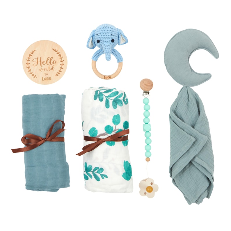 May include: A collection of baby items, including a blue elephant rattle, a wooden teething ring, a muslin swaddle blanket, a pacifier clip, and a wooden disc with the text "Hello world in Luca". The items are in shades of blue and white.