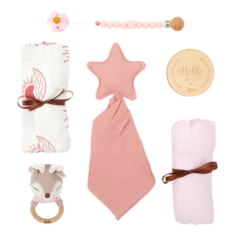 May include: A collection of baby items in shades of pink and white. Includes a pacifier clip, a star-shaped plush, a muslin cloth, a wooden sign with the text "Hello world in Hello", a rolled blanket, and a crocheted deer rattle.