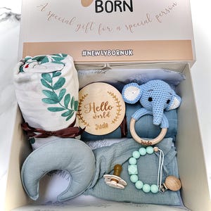 May include: A gift box for a newborn baby, containing a muslin blanket with green leaf print, a wooden teething ring, a blue crochet elephant toy, a pacifier, and a teething necklace. The box is labeled "Newly Born" and "A special gift for a special person."