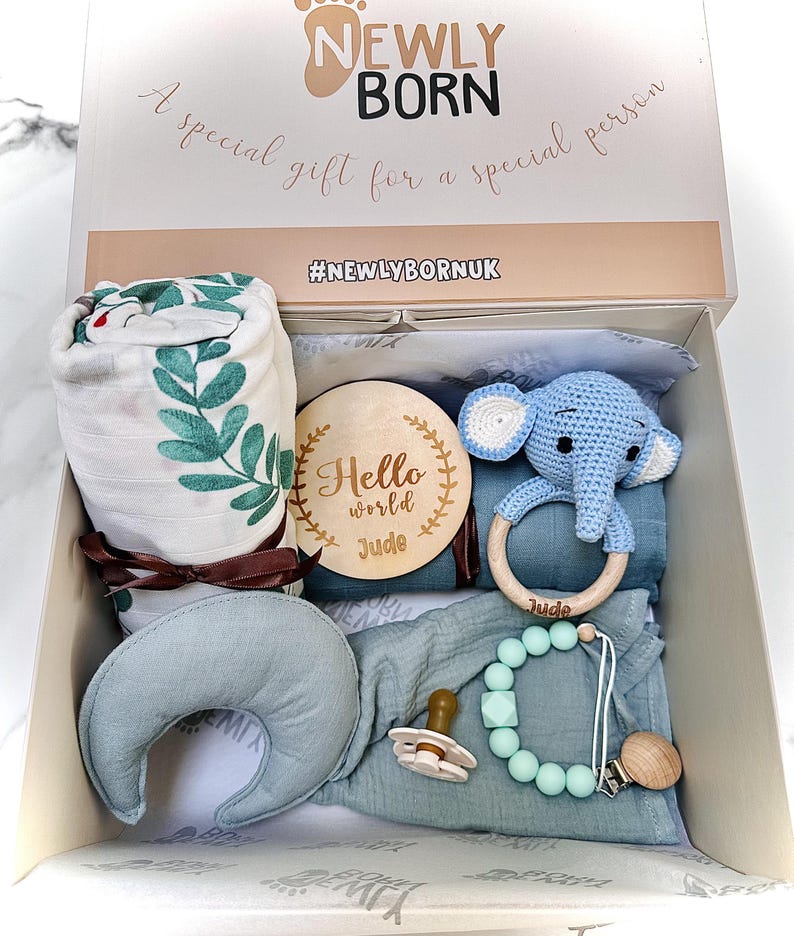 May include: A gift box for a newborn baby, containing a muslin blanket with green leaf print, a wooden teething ring, a blue crochet elephant toy, a pacifier, and a teething necklace. The box is labeled "Newly Born" and "A special gift for a special person."