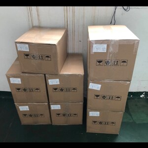Puede incluir: Una pila de nueve cajas de cart&oacute;n marr&oacute;n con flechas negras impresas y un s&iacute;mbolo de paraguas. Las cajas est&aacute;n apiladas en tres filas de tres cajas cada una.