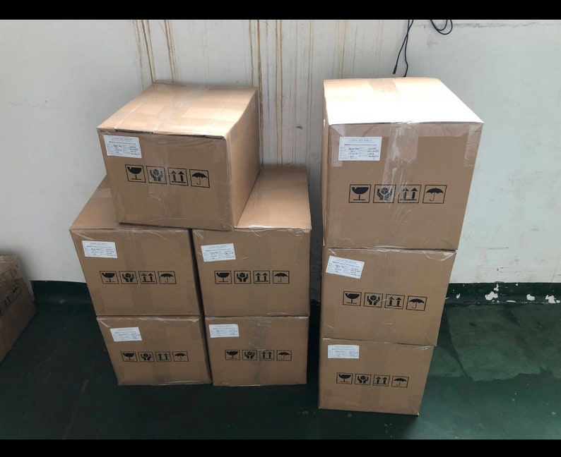 Puede incluir: Una pila de nueve cajas de cart&oacute;n marr&oacute;n con flechas negras impresas y un s&iacute;mbolo de paraguas. Las cajas est&aacute;n apiladas en tres filas de tres cajas cada una.