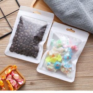 Puede incluir: Dos bolsas de pl&aacute;stico transparentes de pie con bordes blancos y agujeros para colgar. Una bolsa contiene granos de caf&eacute; y la otra contiene caramelos de colores.