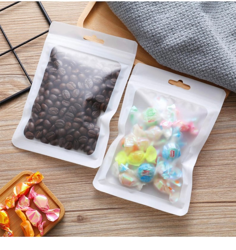 Puede incluir: Dos bolsas de pl&aacute;stico transparentes de pie con bordes blancos y agujeros para colgar. Una bolsa contiene granos de caf&eacute; y la otra contiene caramelos de colores.