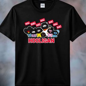 May include: A black short-sleeved t-shirt featuring a graphic of seven colorful cartoon characters wearing black masks. Above the characters are multiple red speech bubbles with the text HA! and the word HOOLIGAN printed in red below them.