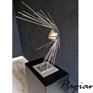 Peut inclure: Une sculpture en métal argenté avec une sphère au sommet et de multiples tiges métalliques fines rayonnant de la sphère. La sculpture est sur une base carrée noire.
