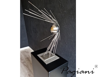 Op de afbeelding: Een zilveren metalen sculptuur met een bol aan de bovenkant en meerdere dunne metalen staven die uit de bol stralen. De sculptuur staat op een zwarte vierkante basis.